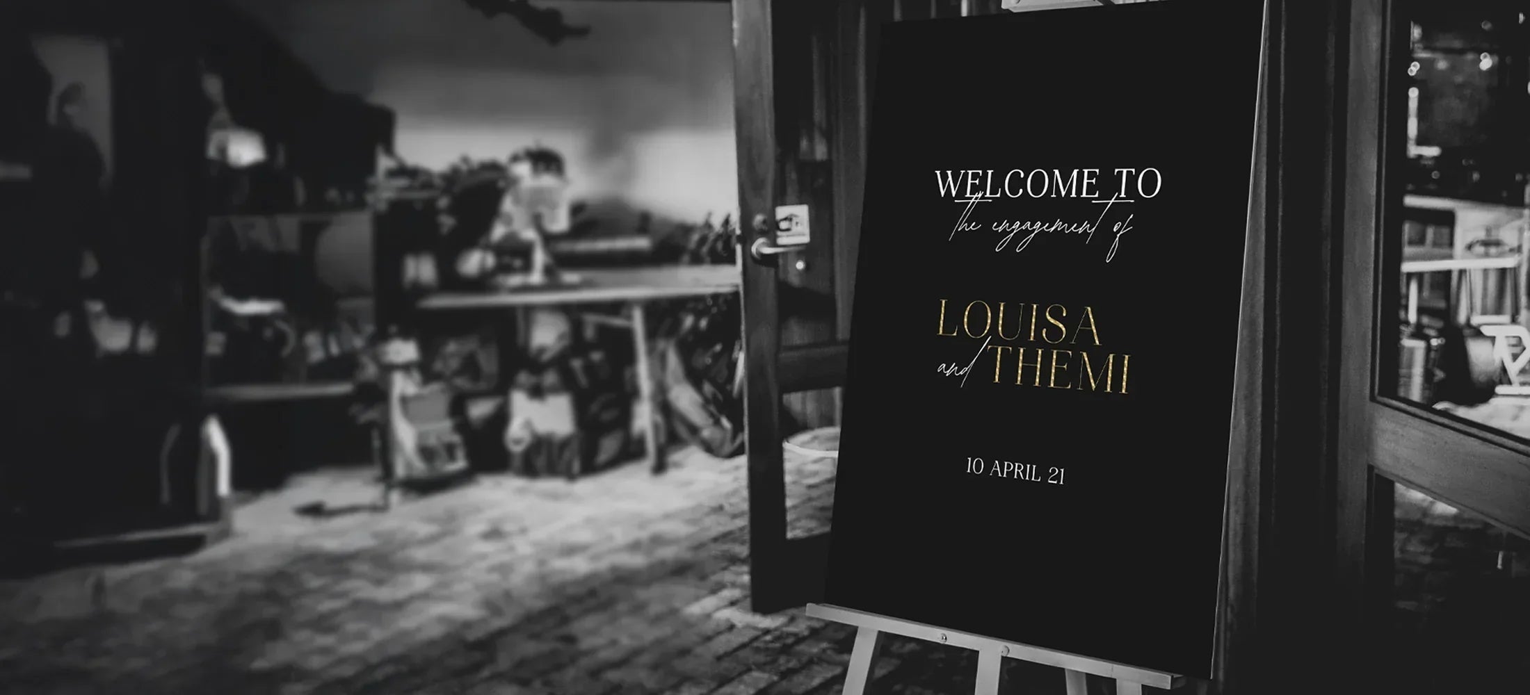 Black engagement welcome sign with gold foil text 'Welcome to the engagement of Louisa and Themi 10 April 21' on wooden easel at evening venue by Felicitations