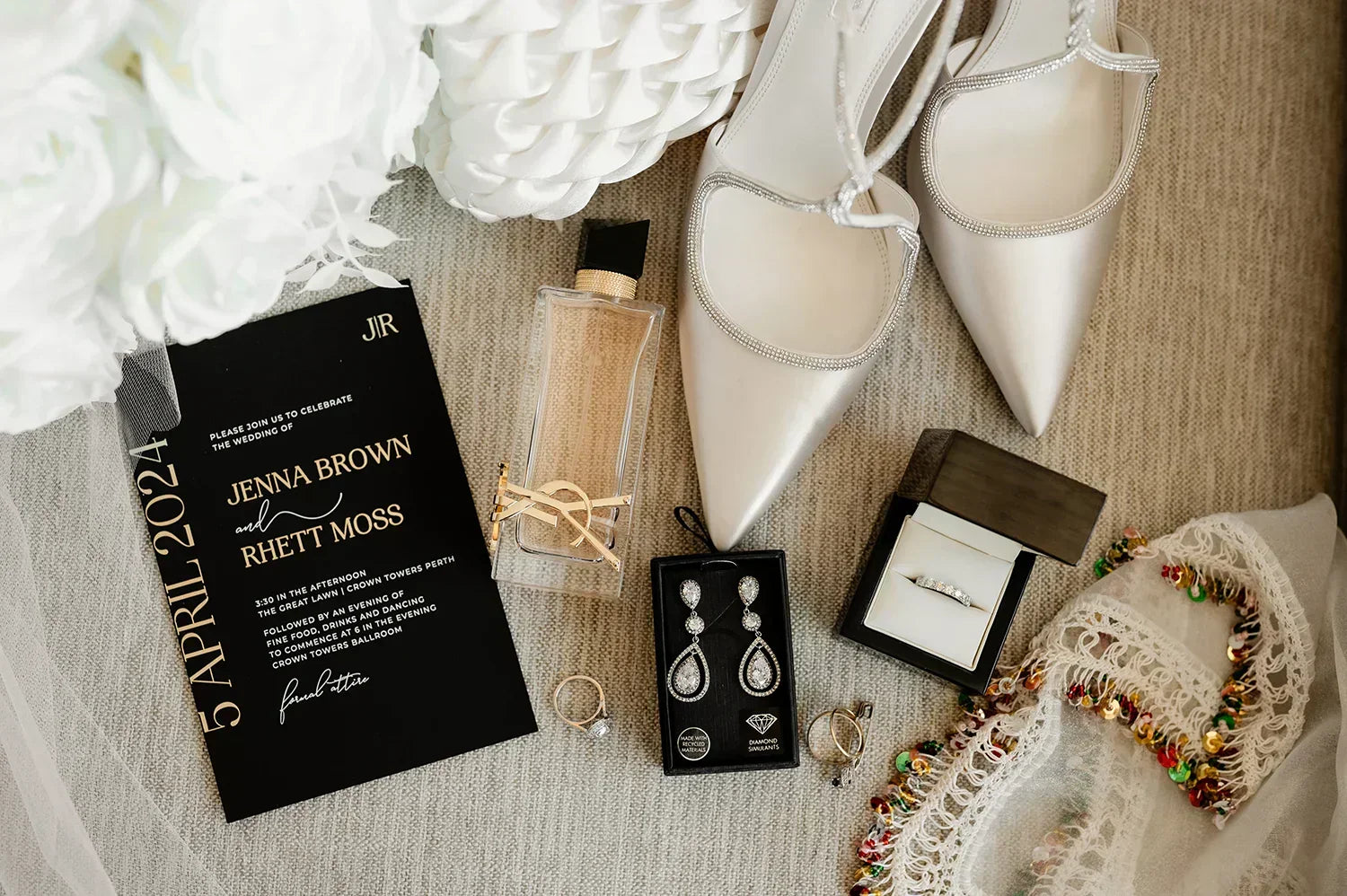 Wedding accessories including white pointed shoes, black and gold wedding invitation, jewelry, perfume, and white floral decorations