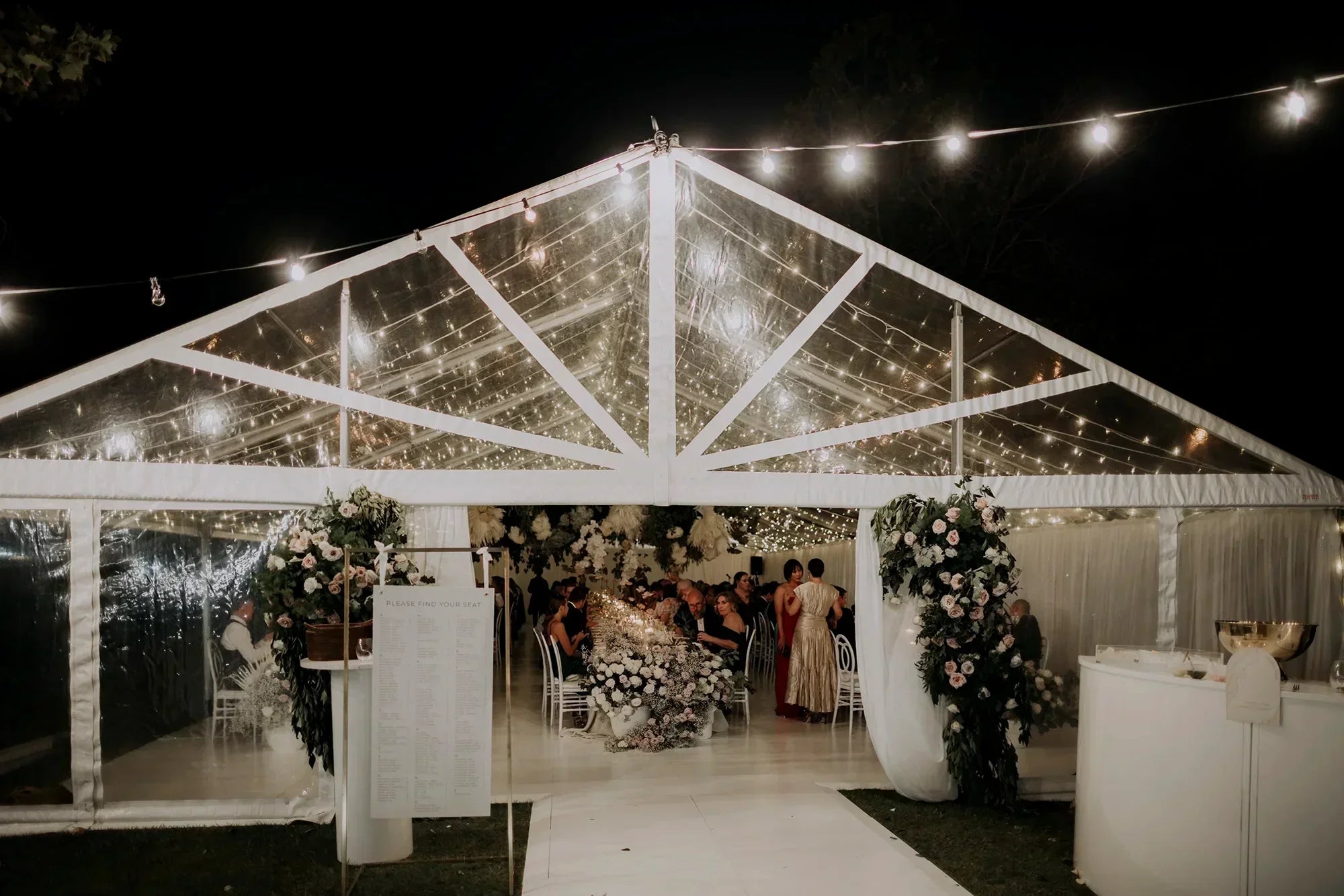 Clear marquee wedding reception at night with fairy lights, white floral arches, custom seating chart sign, and guests dining inside by Felicitations