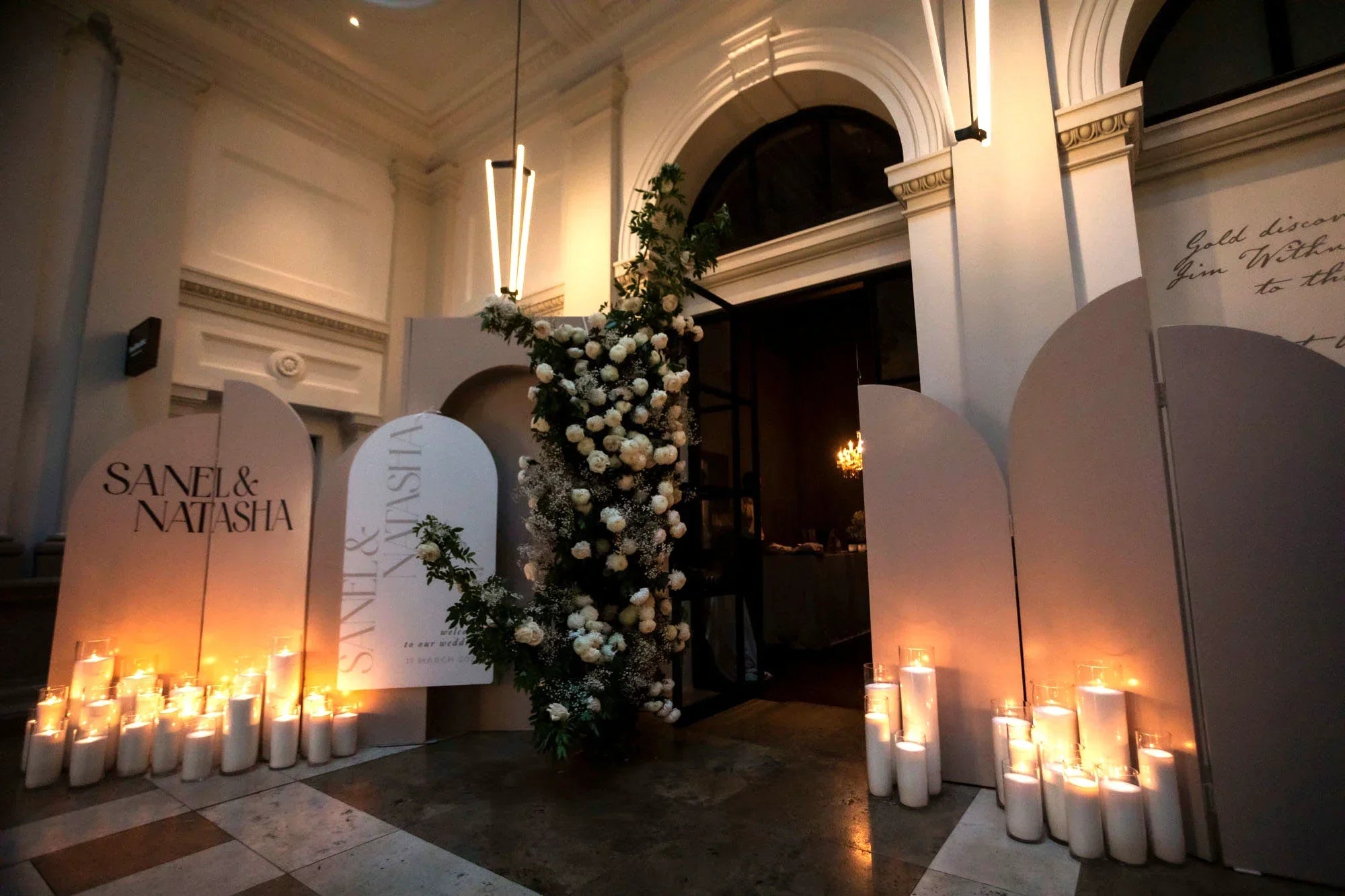 Sanel and Natasha arched wedding welcome sign display with cascading white floral arrangement, pillar candles, and modern lighting at venue entrance by Felicitations