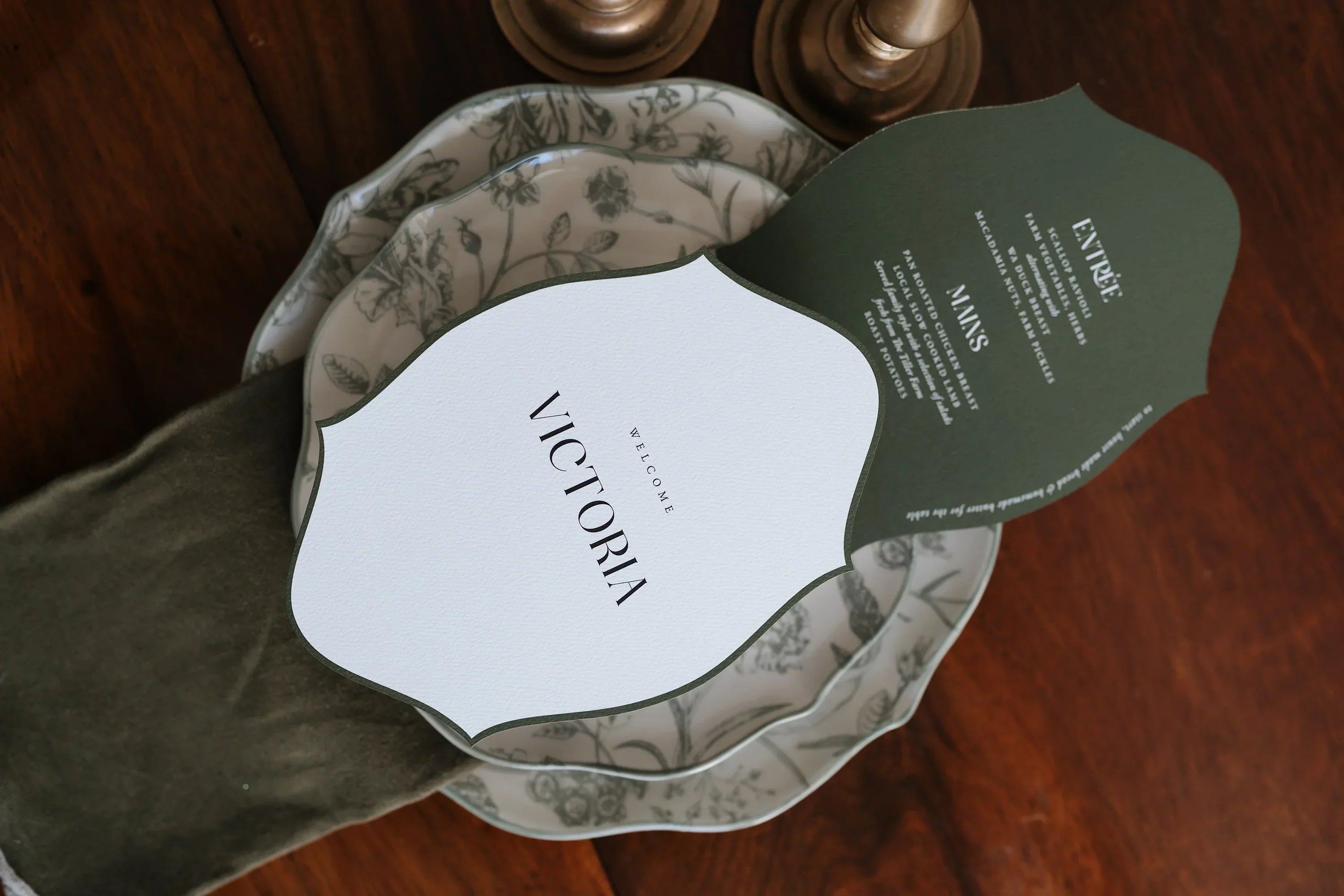 Luxury wedding place card and menu suite for 'Welcome Victoria' featuring olive green and white botanical scalloped design layered on a patterned plate by Felicitations