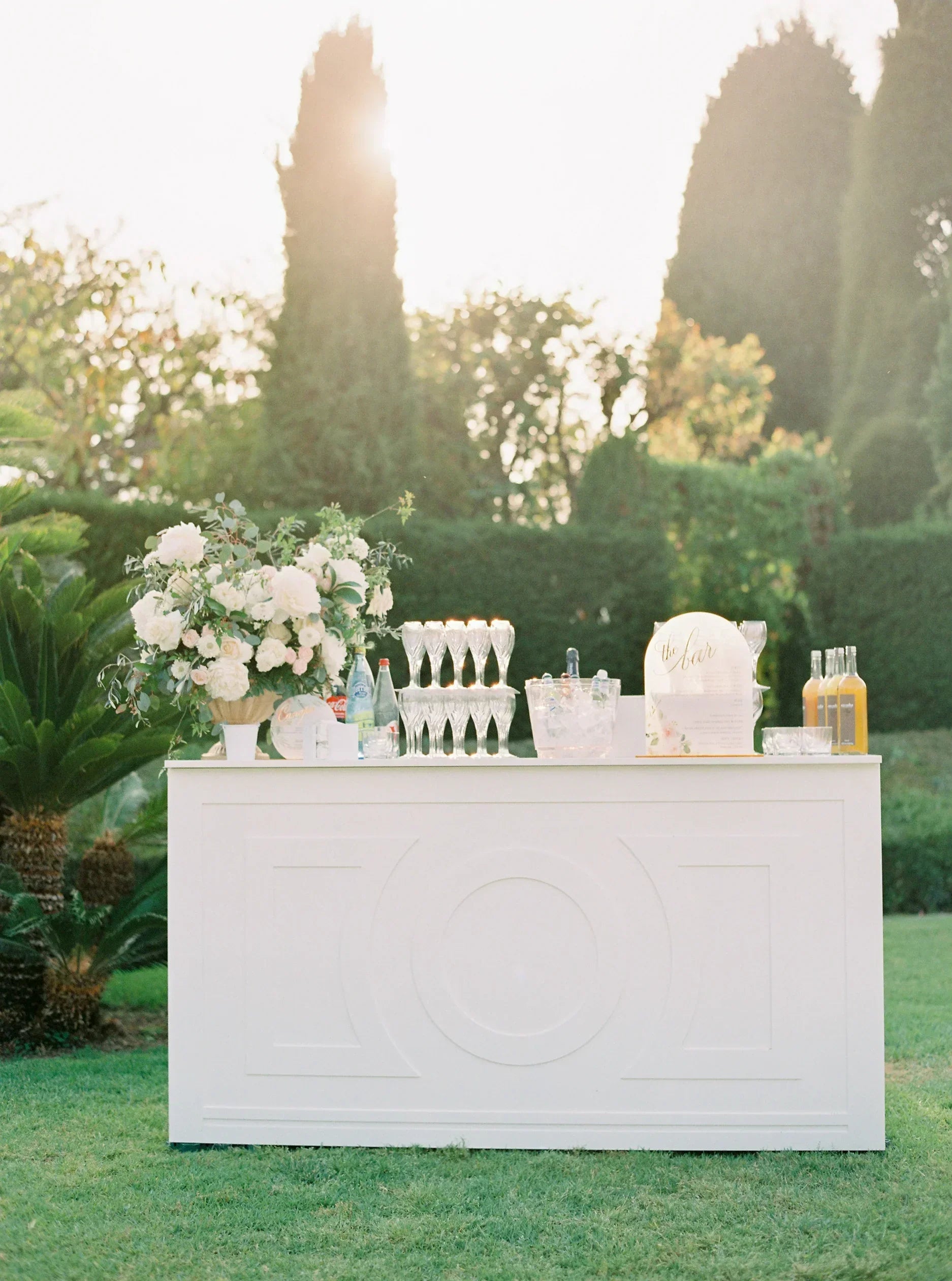 Elegant white wedding bar with white peony floral arrangement, champagne flutes, ice bucket, custom round signage, and drinks in sunlit garden setting by Felicitations