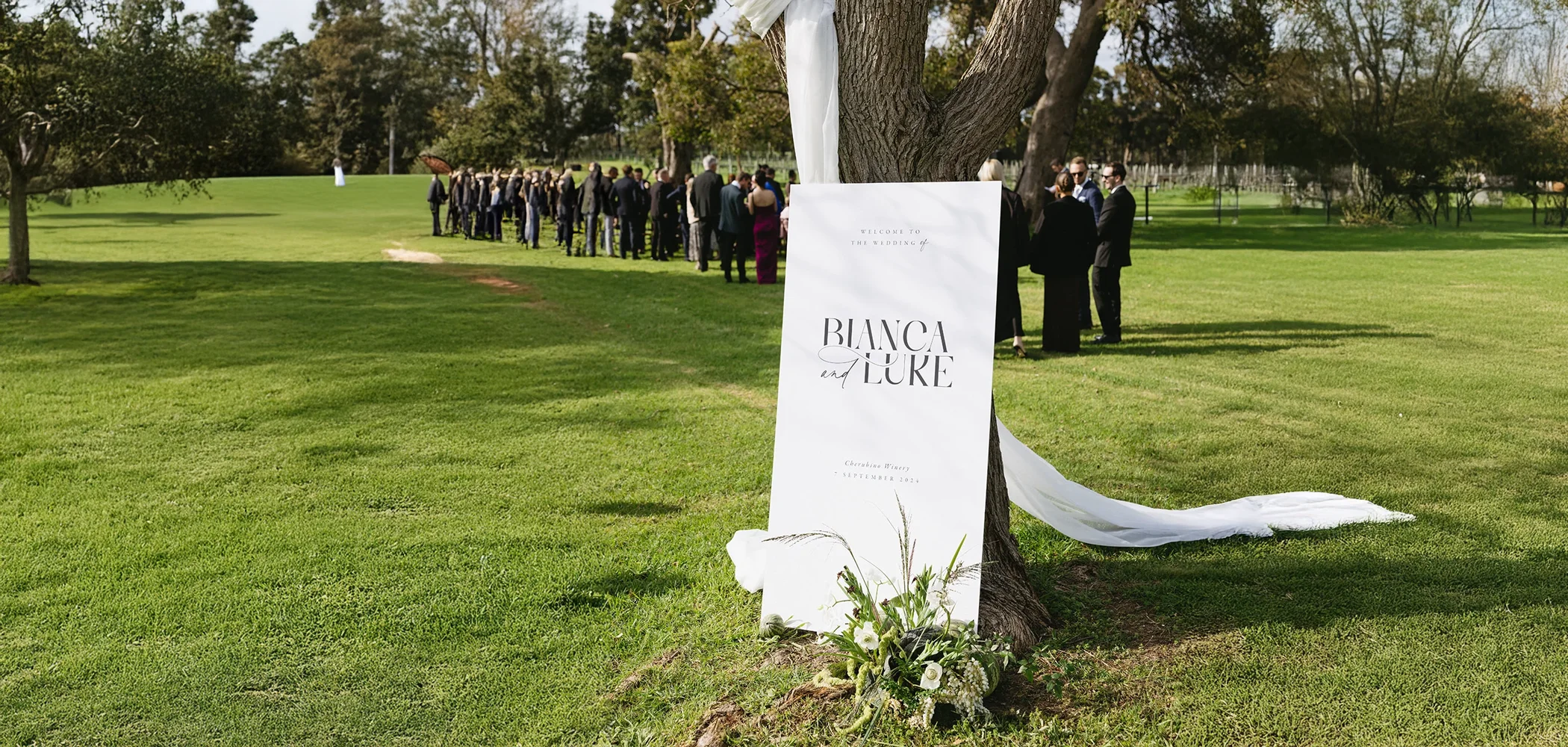 A large white wedding welcome sign for 'Bianca and Luke', displayed against a tree for an elegant outdoor park ceremony.