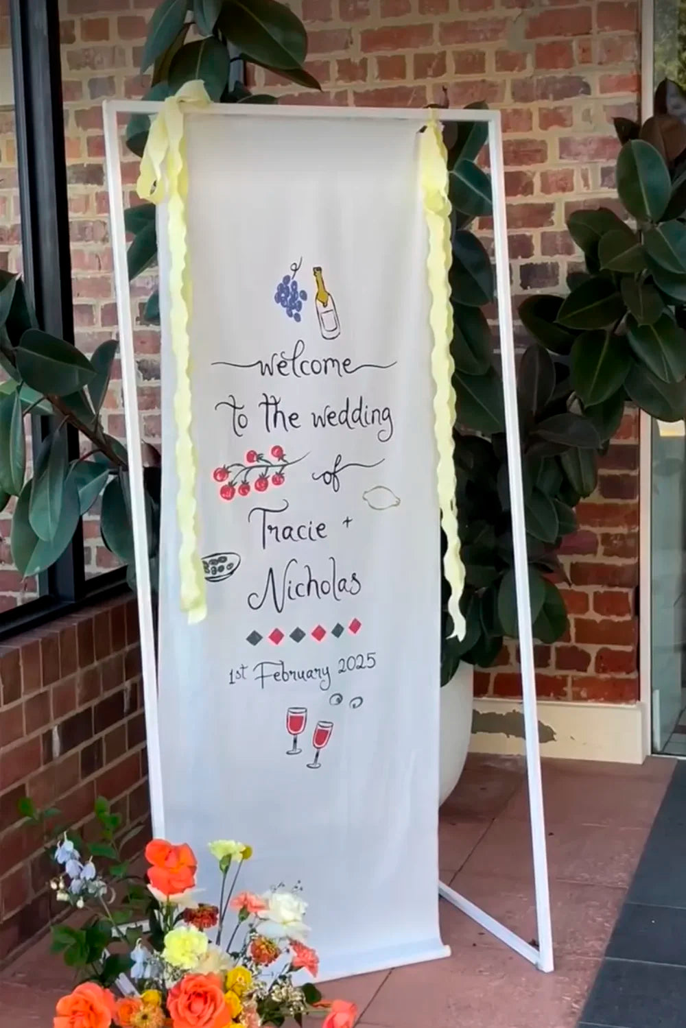 A welcome sign printed on a white fabric banner, featuring charming, colourful illustrations of food and wine for a relaxed wedding celebration.