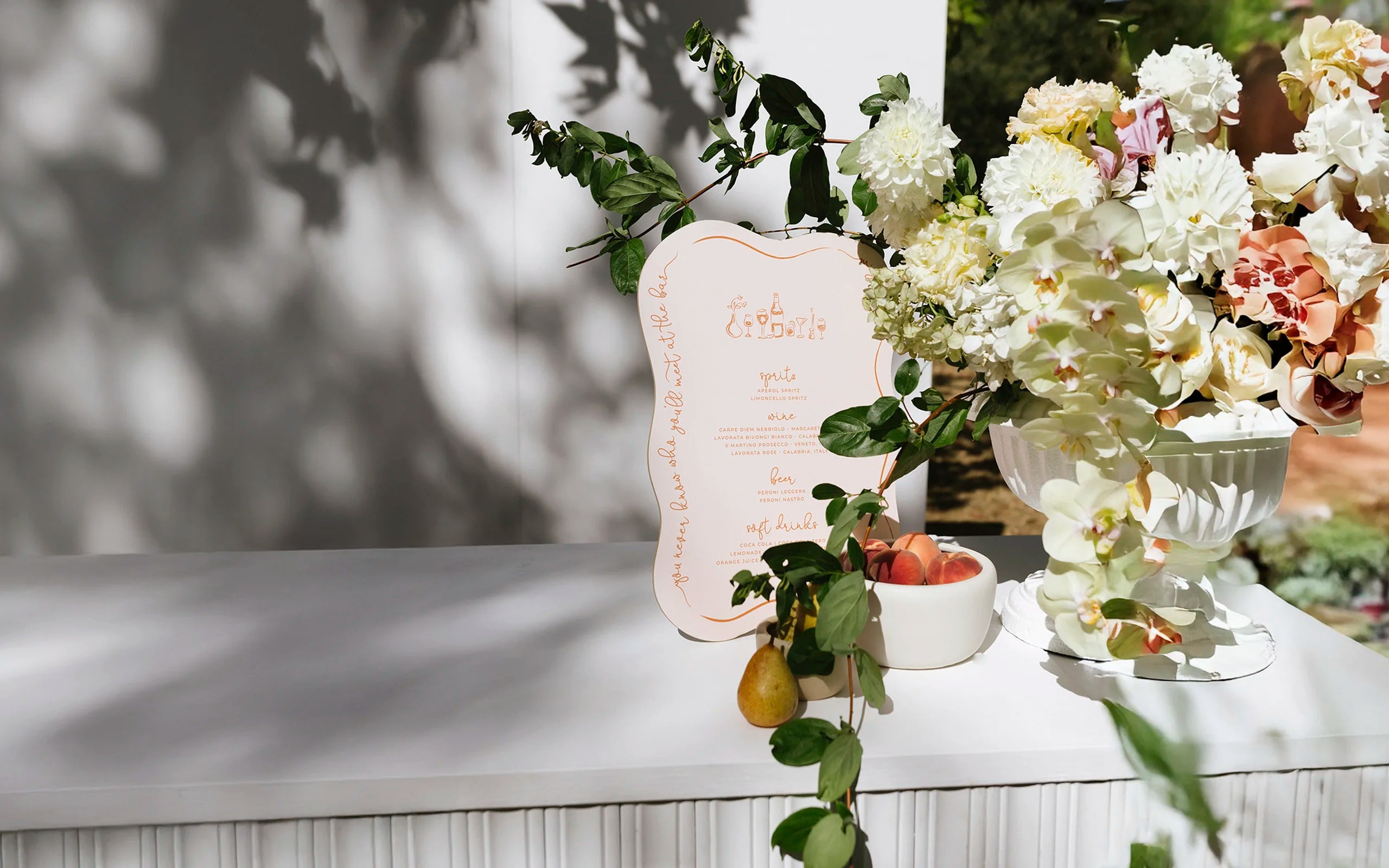 Elegant wedding bar sign by Felicitations Wedding Stationery, styled with a peach-toned drinks menu, fresh fruit, and lush floral arrangement in an outdoor setting.