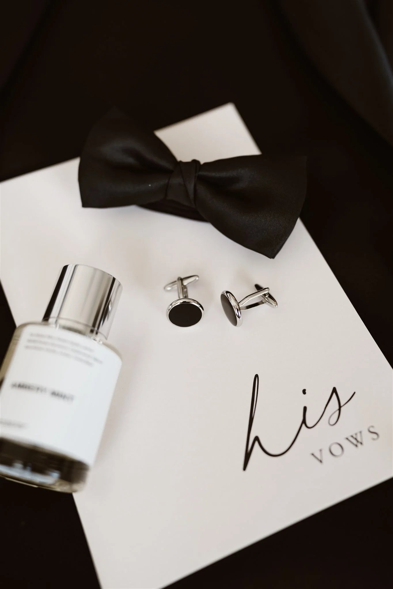 A "His Vows" booklet styled in a flat-lay with groom's accessories including a black bow tie, cufflinks, and cologne.