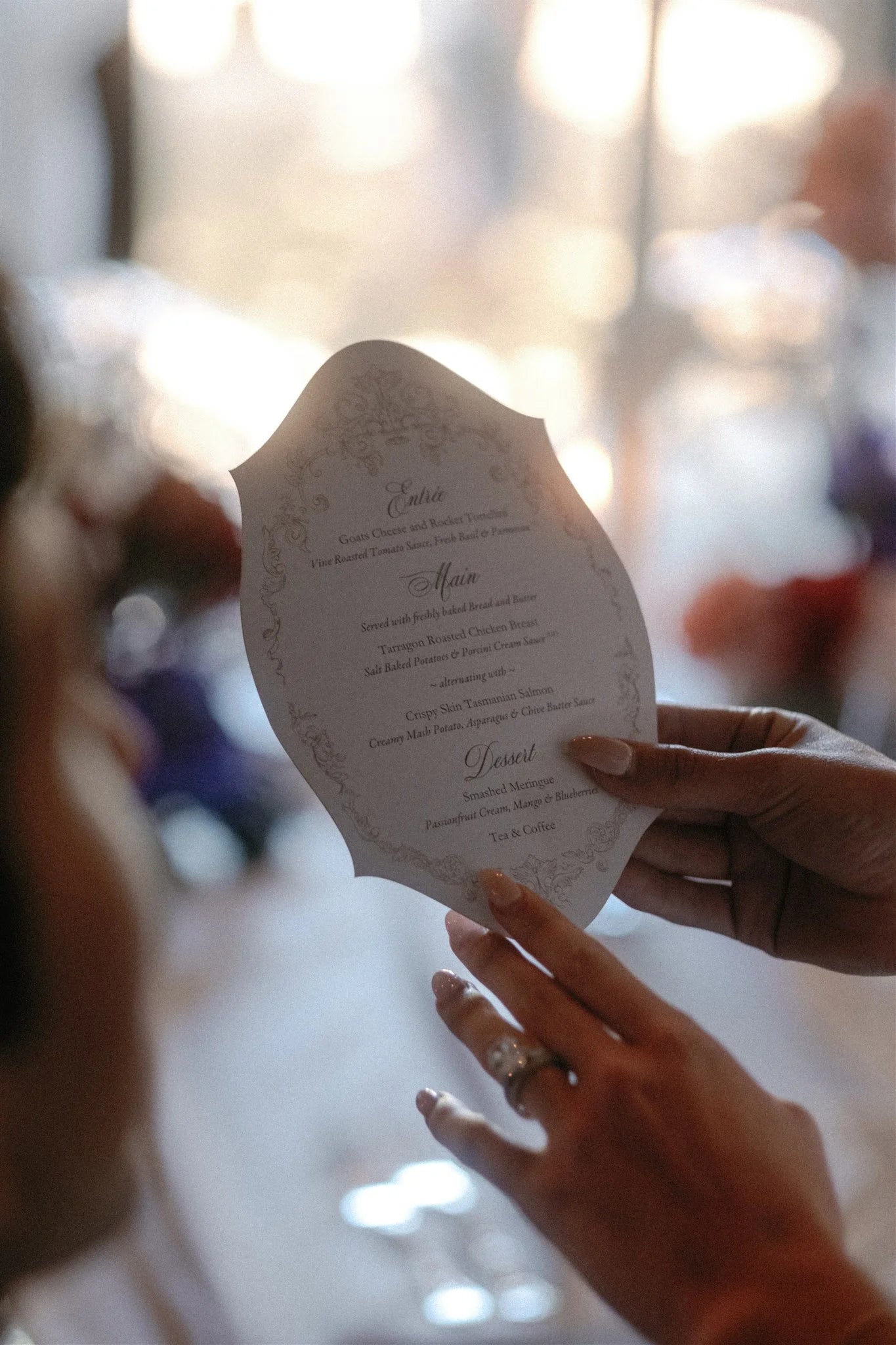 A wedding guest holding an elegant, custom die-cut menu with an ornate border and classic typography at a reception.
