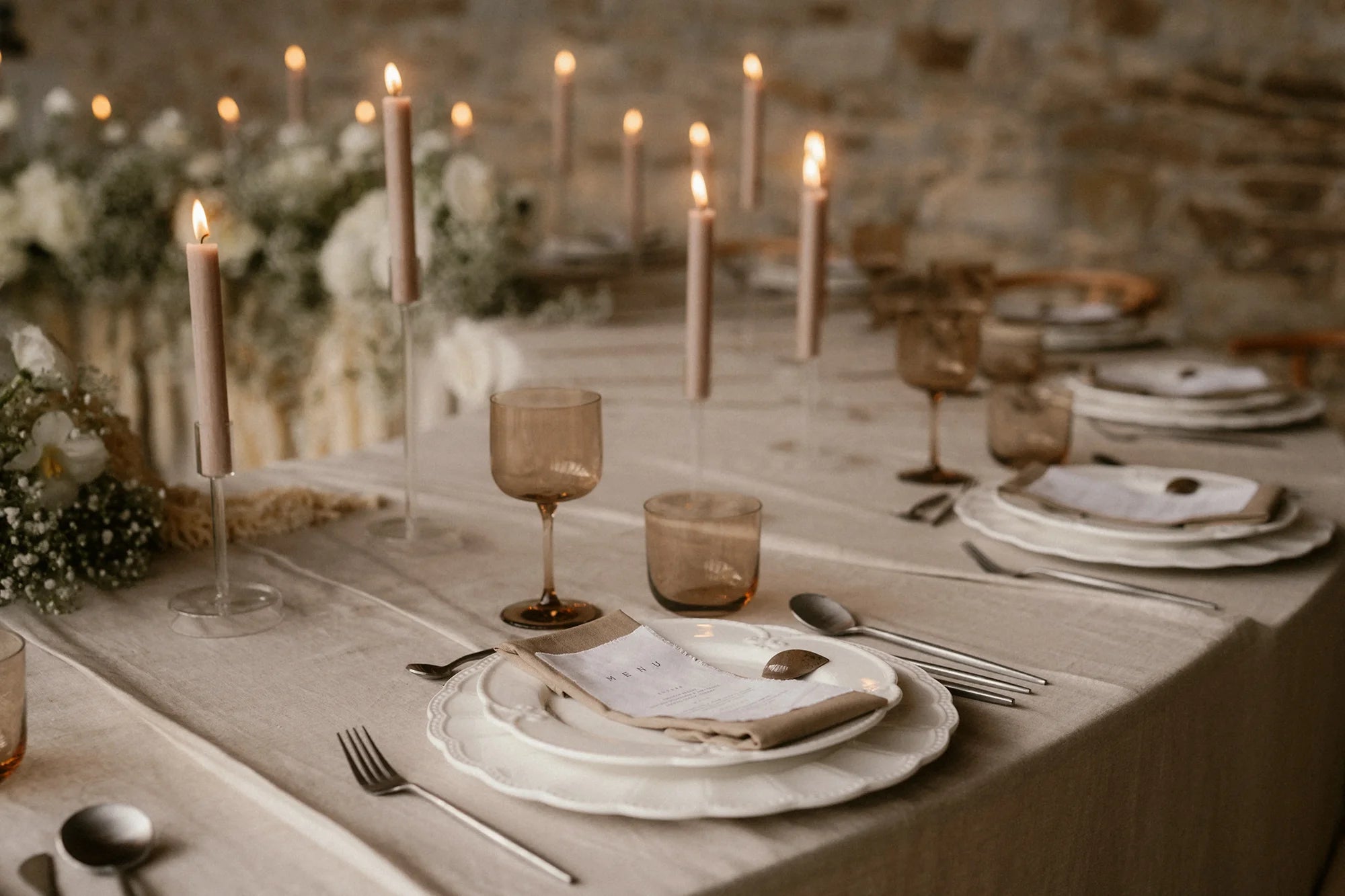A romantic, candlelit wedding tablescape featuring linen menus on ornate plates, amber glassware, and dozens of glowing taper candles.