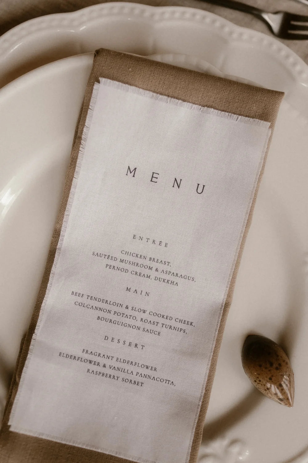 An elegant wedding place setting featuring a custom guest menu printed on fine linen with delicate frayed edges, placed in a folded napkin.