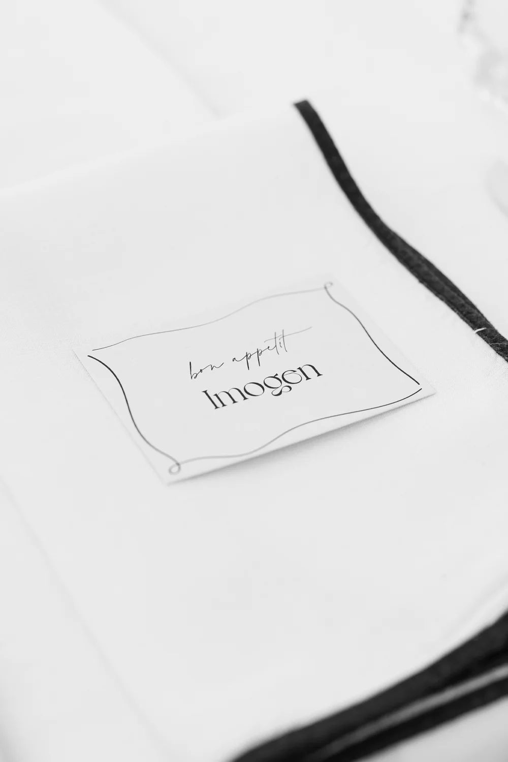 A minimalist wedding place card with the name 'Imogen' in an elegant script, featuring a simple 'bon appetit' message and a delicate hand-drawn border.