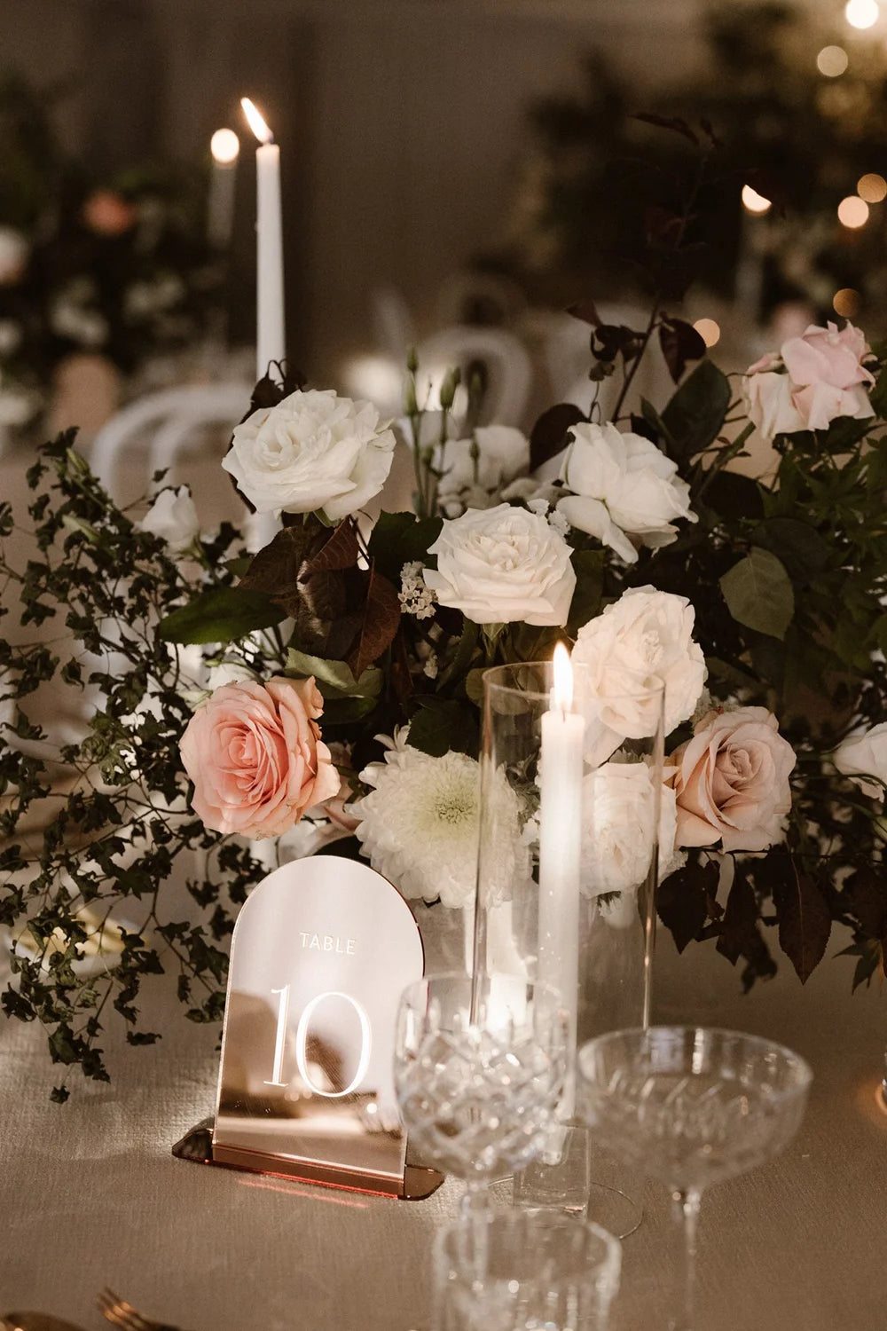 A rose gold mirror acrylic table number with an elegant arched shape, displayed within a lush centerpiece of white and pink roses at a wedding reception.