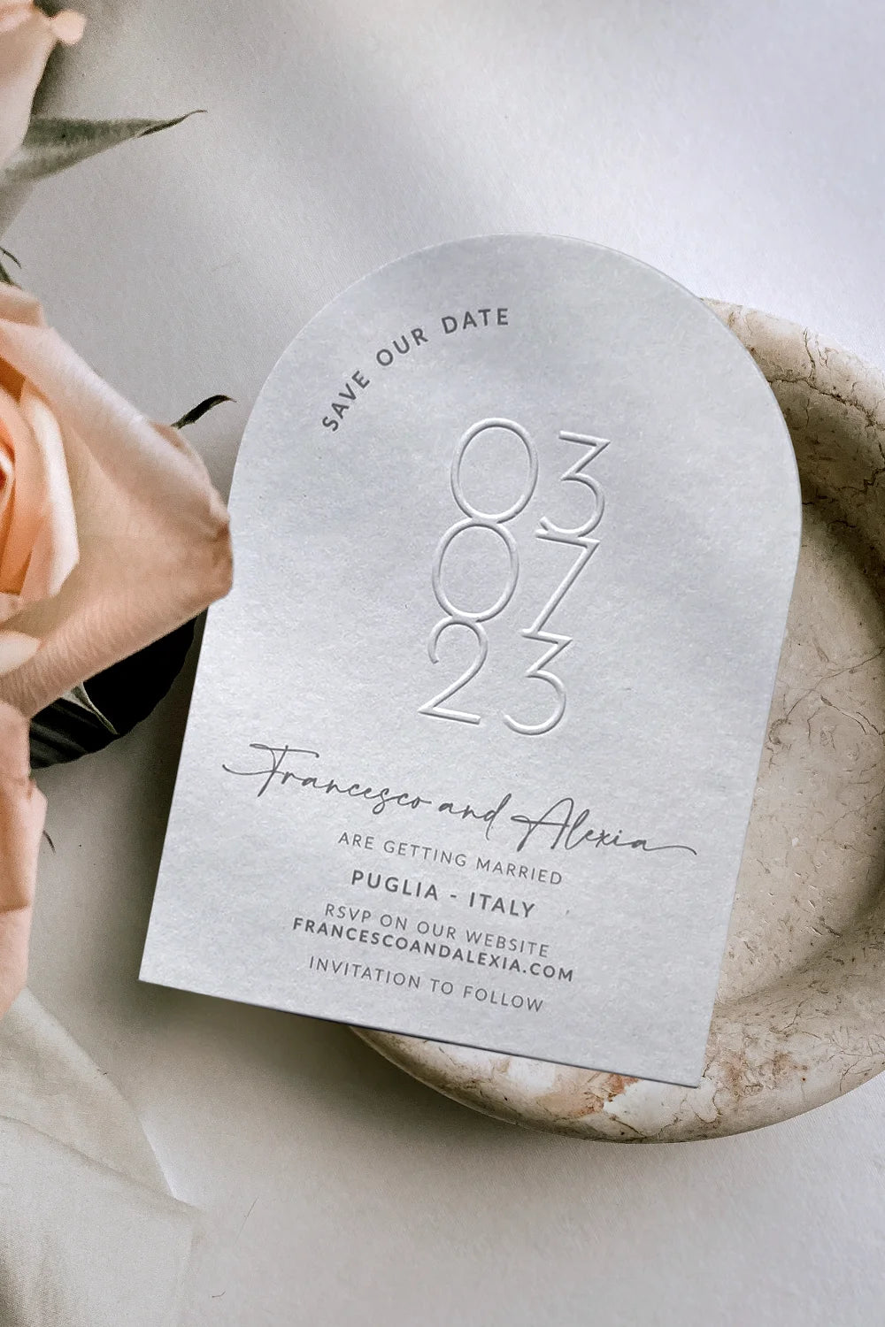 Elegant, arch-shaped Save-the-Date card featuring embossed minimalist date numerals, contemporary calligraphy names, and sophisticated typography details, styled alongside delicate blush roses on a neutral marble background.
