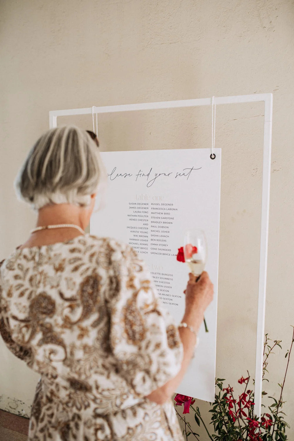 A wedding guest enjoys a glass of champagne while reading the guest list on a minimalist seating chart hanging from a white frame.