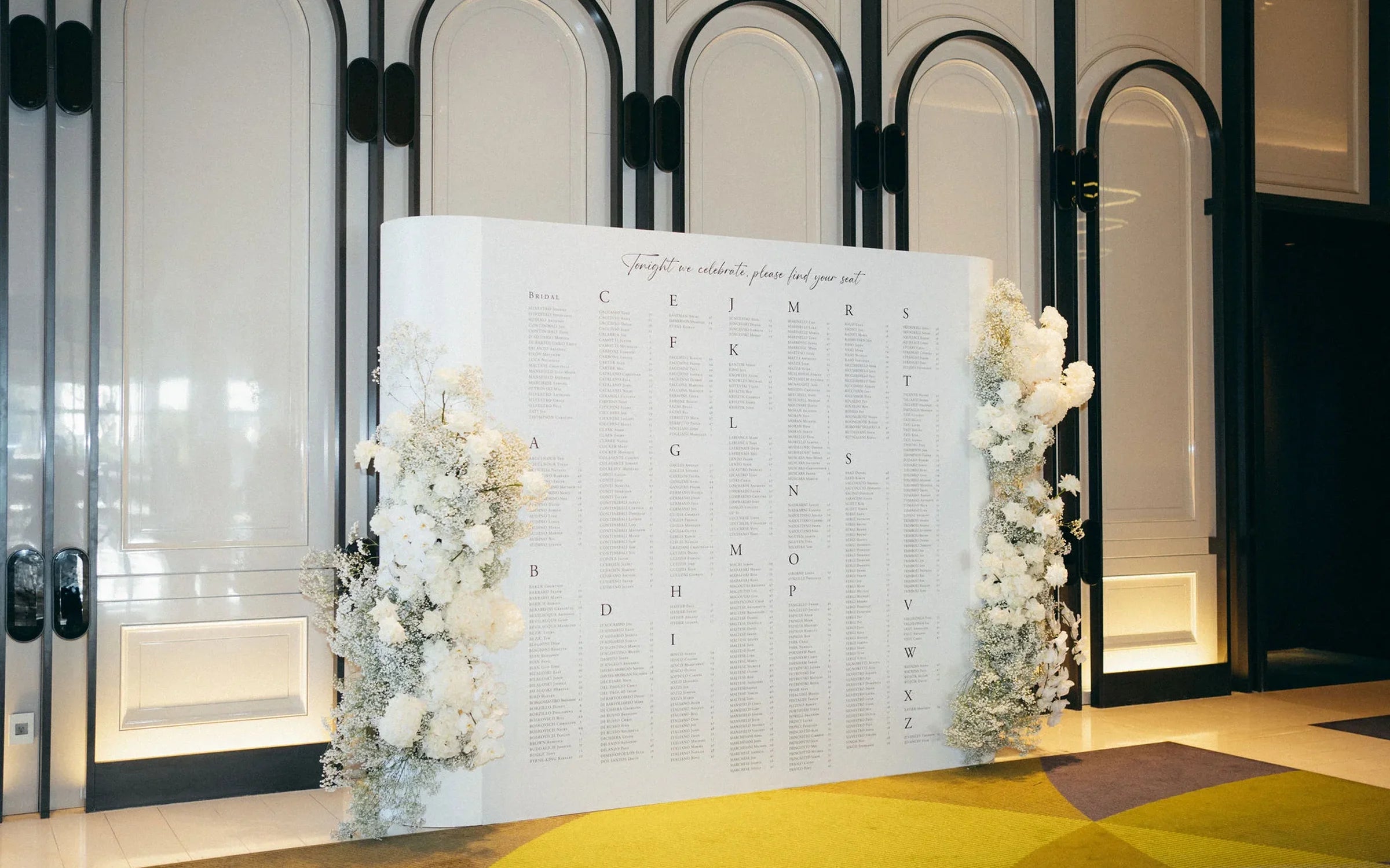 A large, minimalist white wedding seating chart displayed in a modern venue, flanked by stunning floor-standing white floral arrangements.