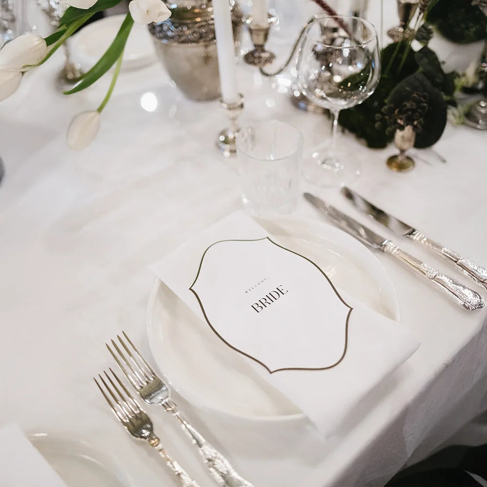 Custom-shaped WELCOME BRIDE place card on white plate with silver cutlery, white tulips and crystal glasses on luxe wedding head table