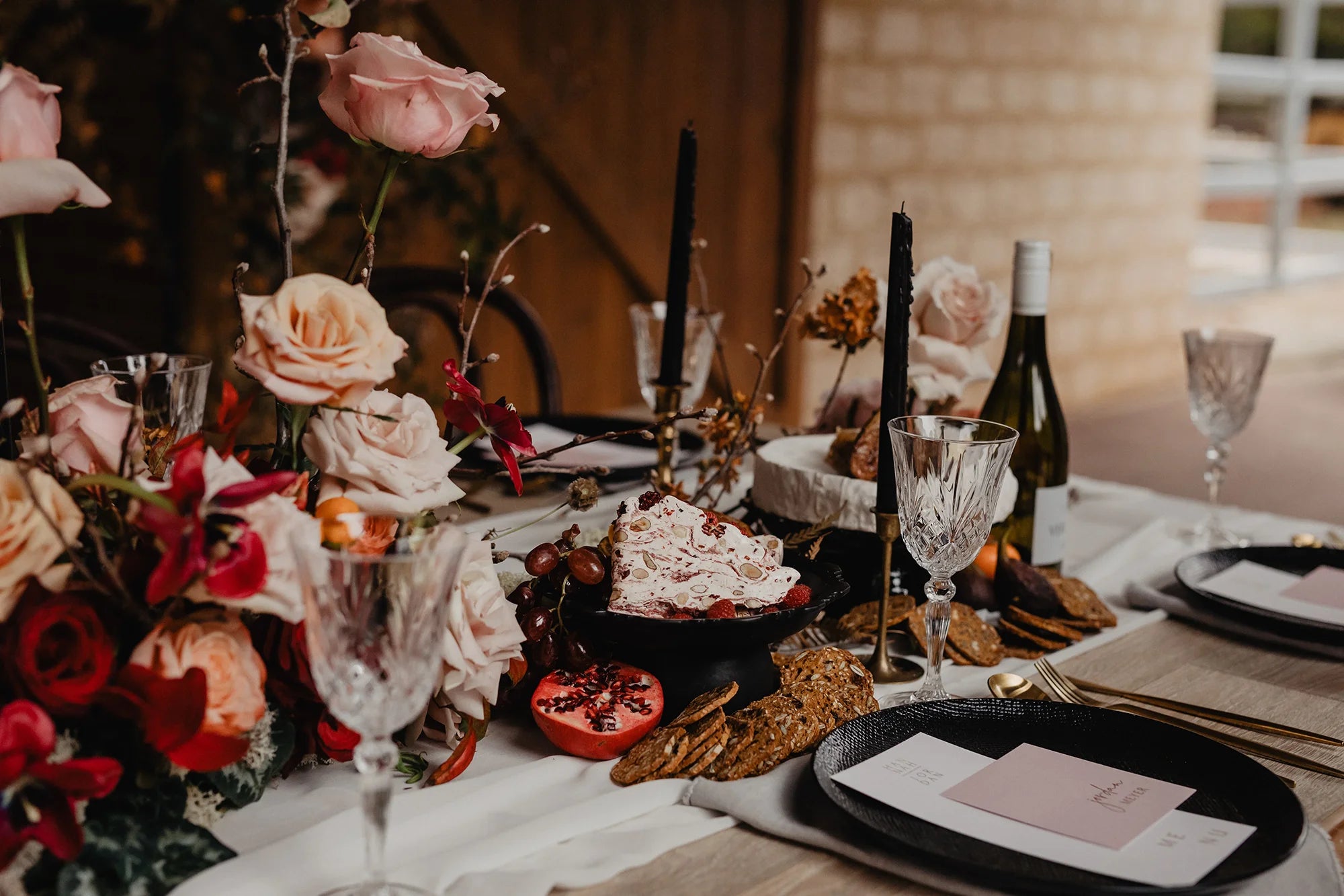 A moody and romantic wedding tablescape featuring a pink guest place card on a black plate, surrounded by blush roses and black taper candles.