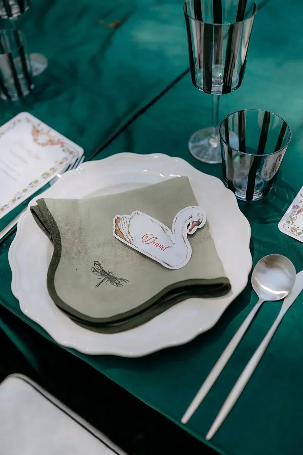 Elegant wedding table setting with white scalloped plate, green napkin, swan place card reading David, silverware, and black-striped glassware on teal tablecloth