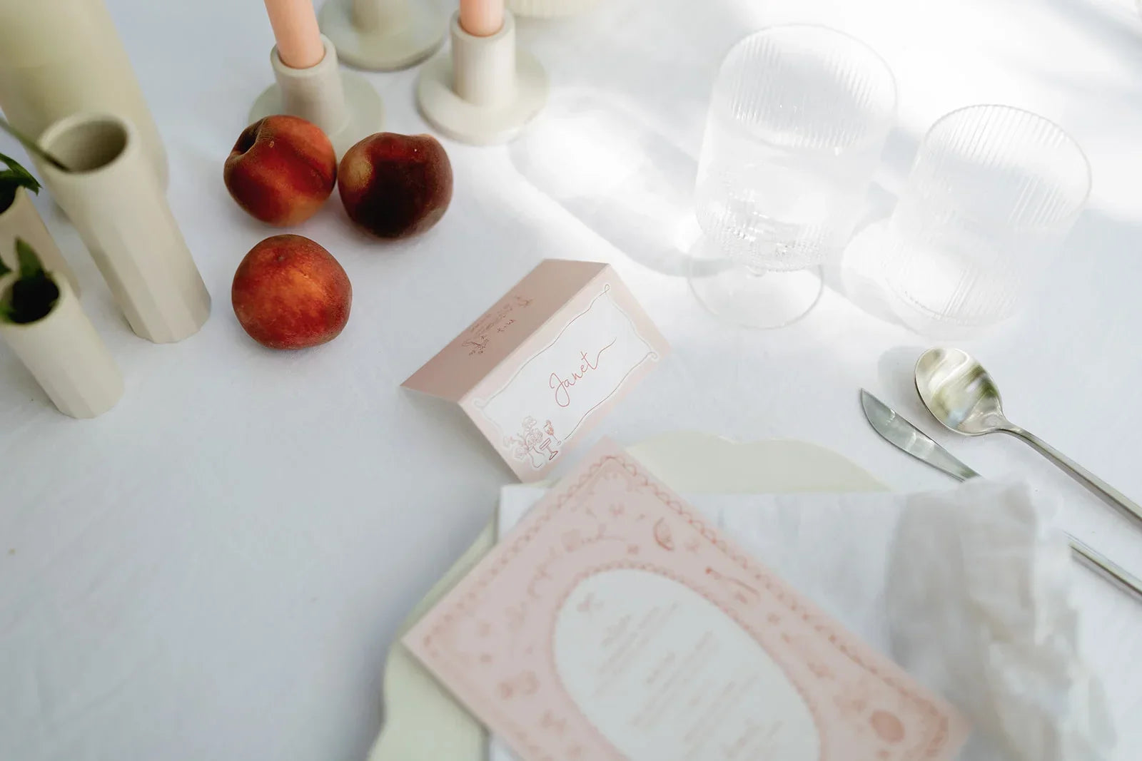 Wedding table setup with folded pink place card labeled Janet, clear glasses, peaches, and cutlery on white linen