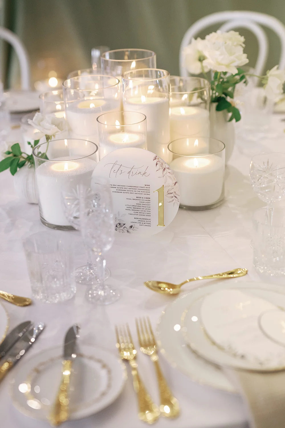 Round wedding drinks menu with gold foil table number 1, surrounded by pillar candles and white roses on luxe reception table