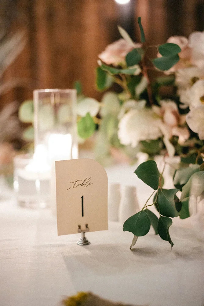 Arch-shaped cardstock wedding table number 1 with calligraphy on silver stand, styled with eucalyptus and white roses at evening reception