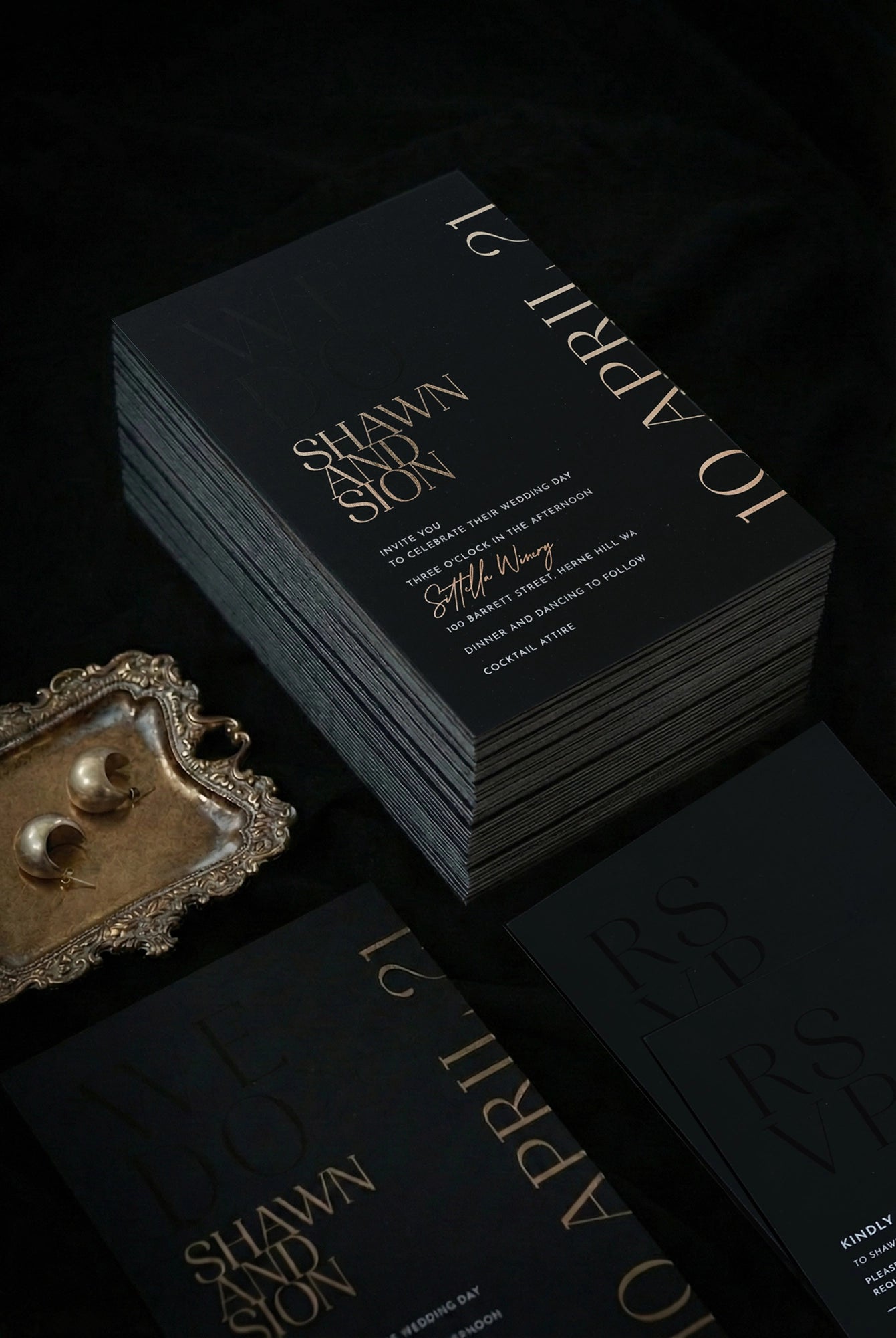 Stack of black Monaco wedding invitations with rose gold foil for Shawn and Sion, ornate vintage tray with gold half-moon earrings beside