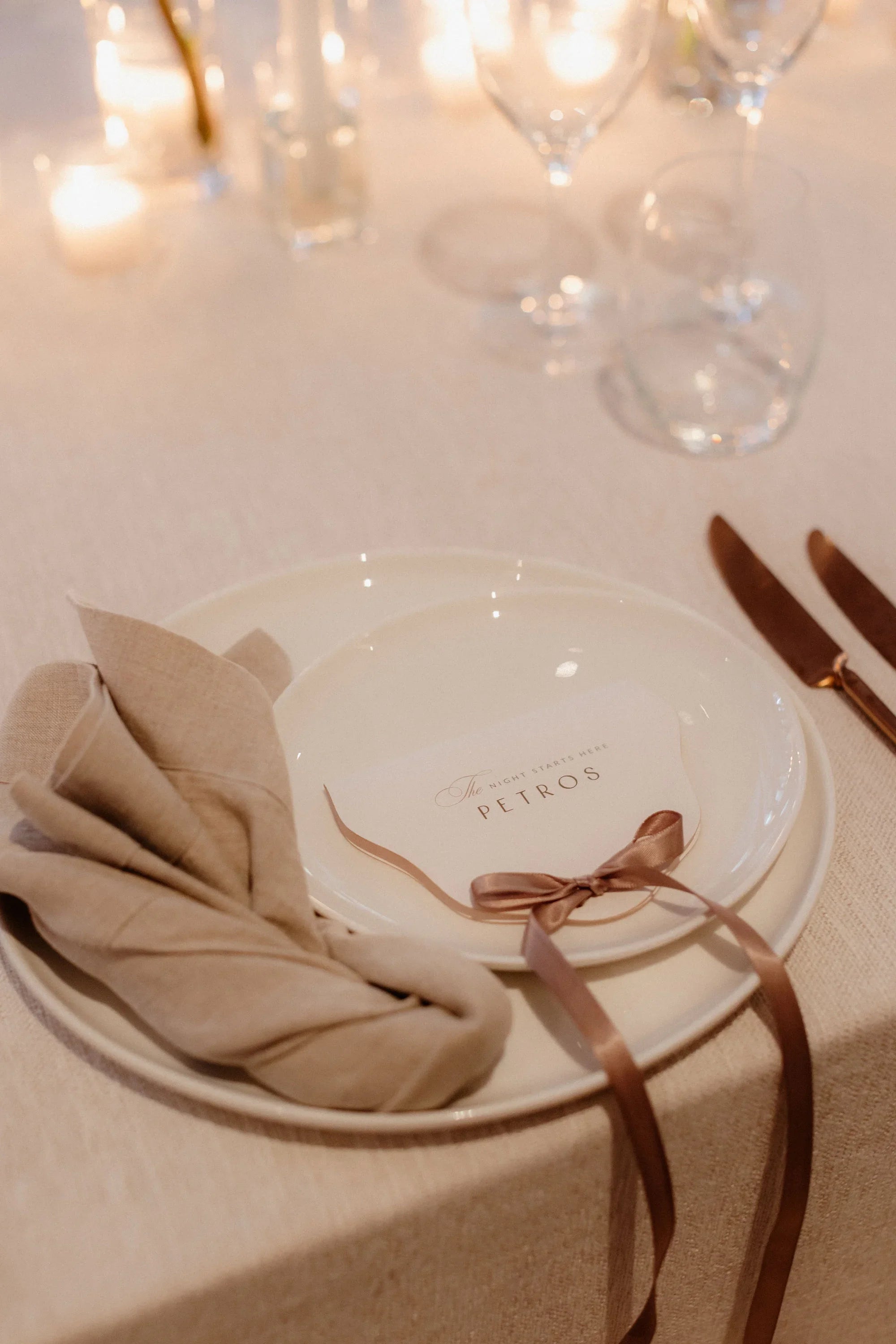 An elegant wedding table setting featuring a round guest place card for 'Petros', finished with a dusty rose silk ribbon bow on a ceramic plate.