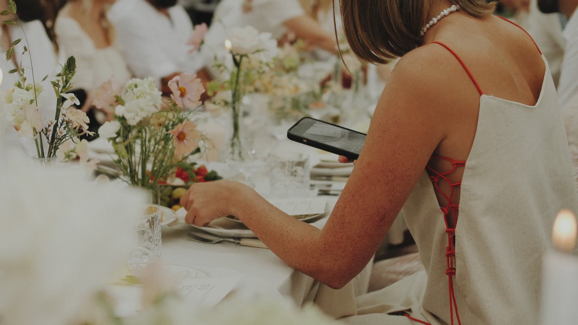 Load video: Close-up of a woman photographing a wedding menu on an elegantly set table with pastel flowers in a greenhouse setting.