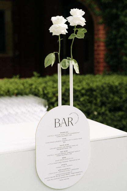 A unique oval-shaped wedding bar menu that doubles as a vase for two white roses, featuring a list of premium local Western Australian wines.
