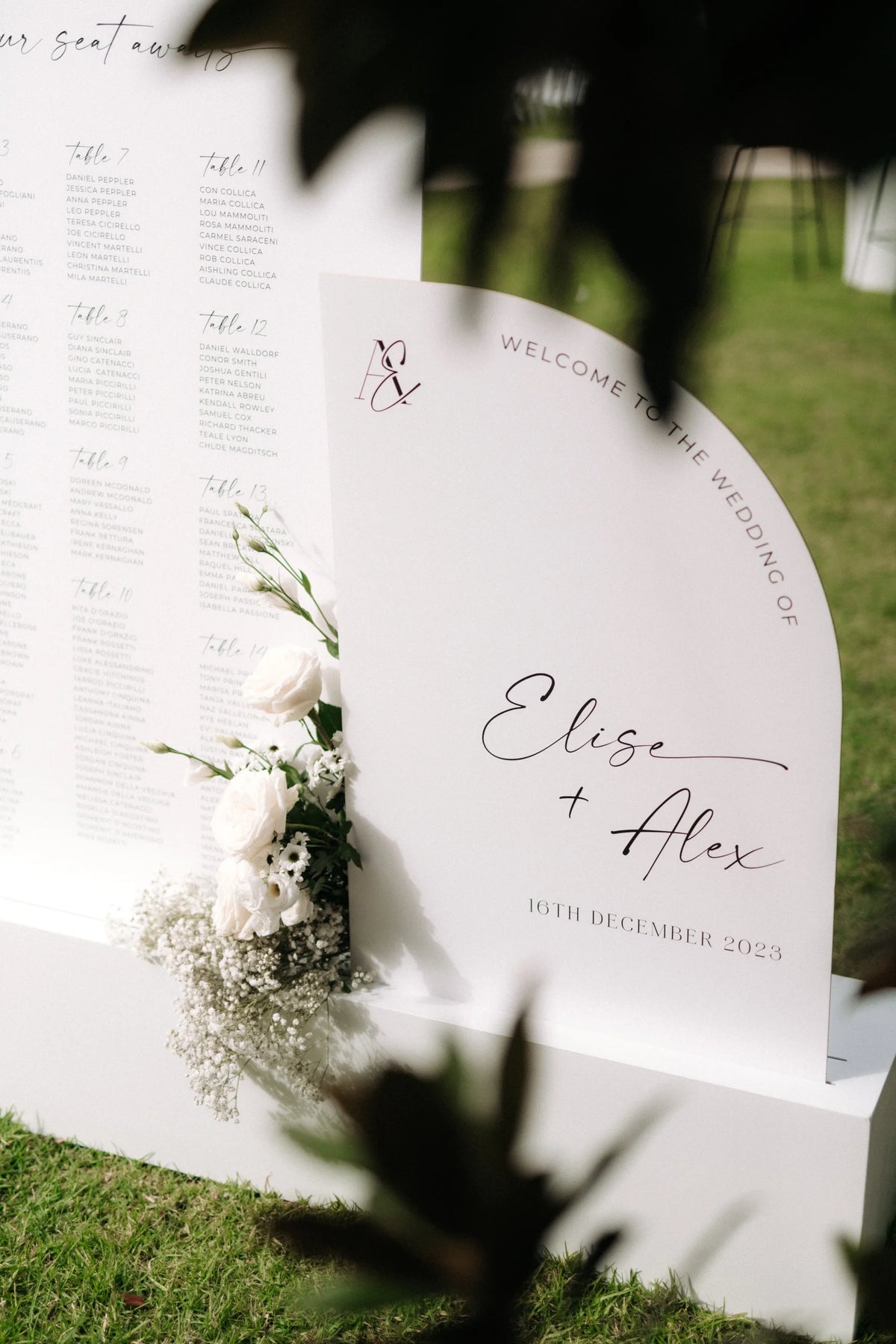 A modern arched wedding welcome sign with a custom monogram, displayed next to a matching seating chart and styled with white roses.