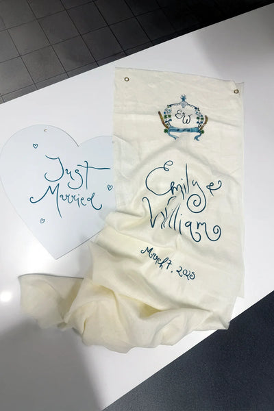 Hand-painted fabric wedding welcome banner for Emily and William with custom blue monogram crest, paired with heart-shaped Just Married sign