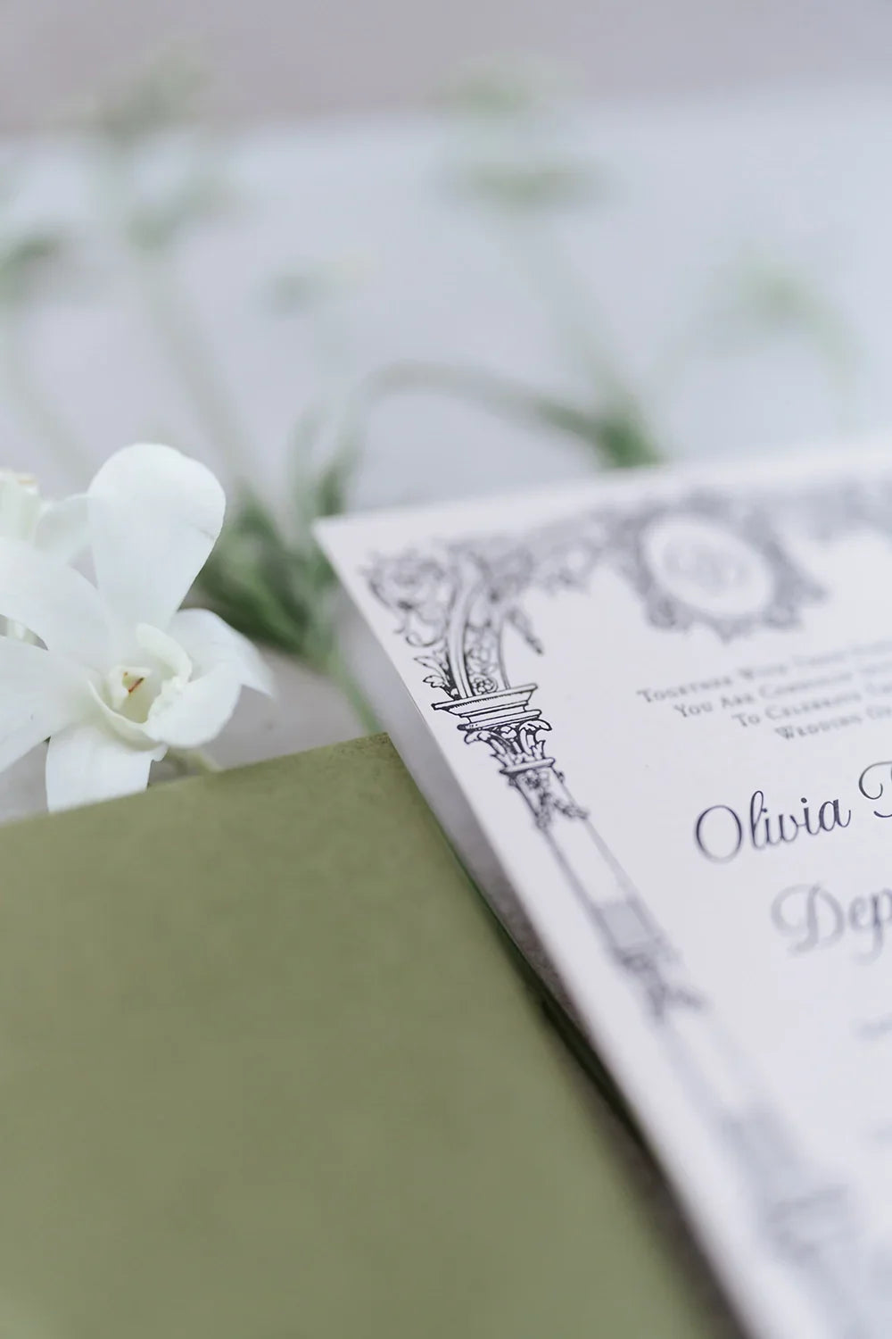 Close-up of elegant wedding invitation card with intricate black border design, white flower, and green envelope