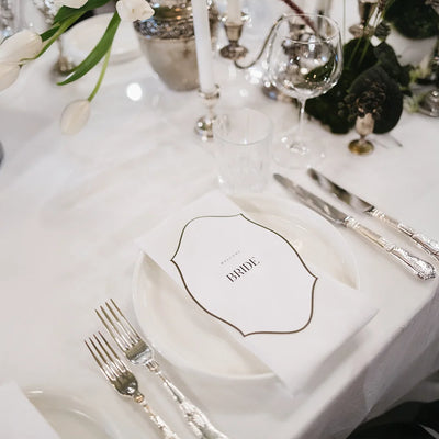 Custom-shaped WELCOME BRIDE place card on white plate with silver cutlery, white tulips and crystal glasses on luxe wedding head table