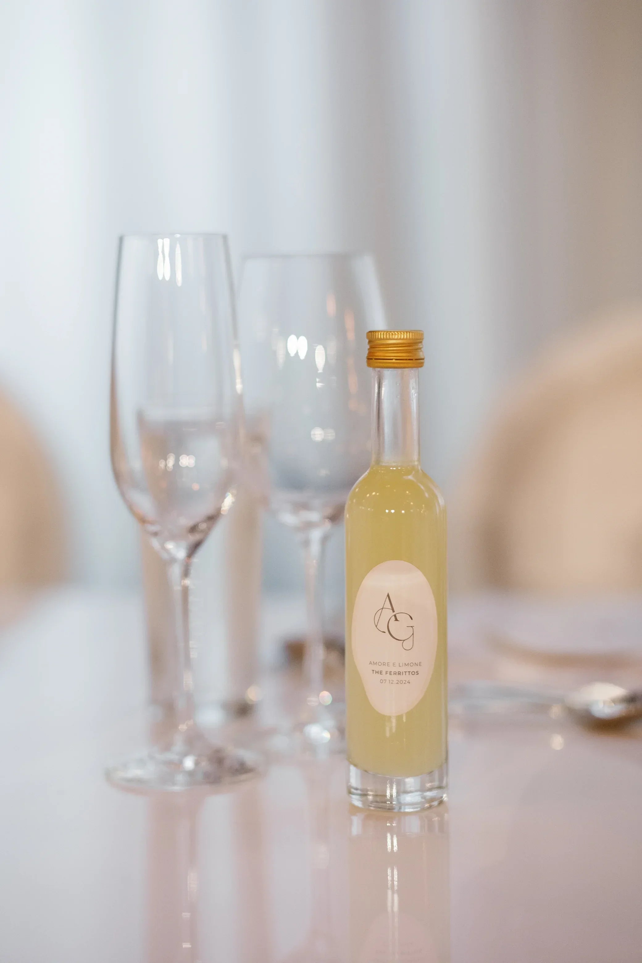 Small glass bottle with yellow liquid and gold cap on a white table with two wine glasses
