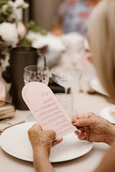 Blush arch shaped wedding menu held at reception table setting by Felicitations Wedding Stationery