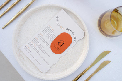 Elegant wedding menu card with scalloped edges and orange tag on speckled plate with gold cutlery and amber glass