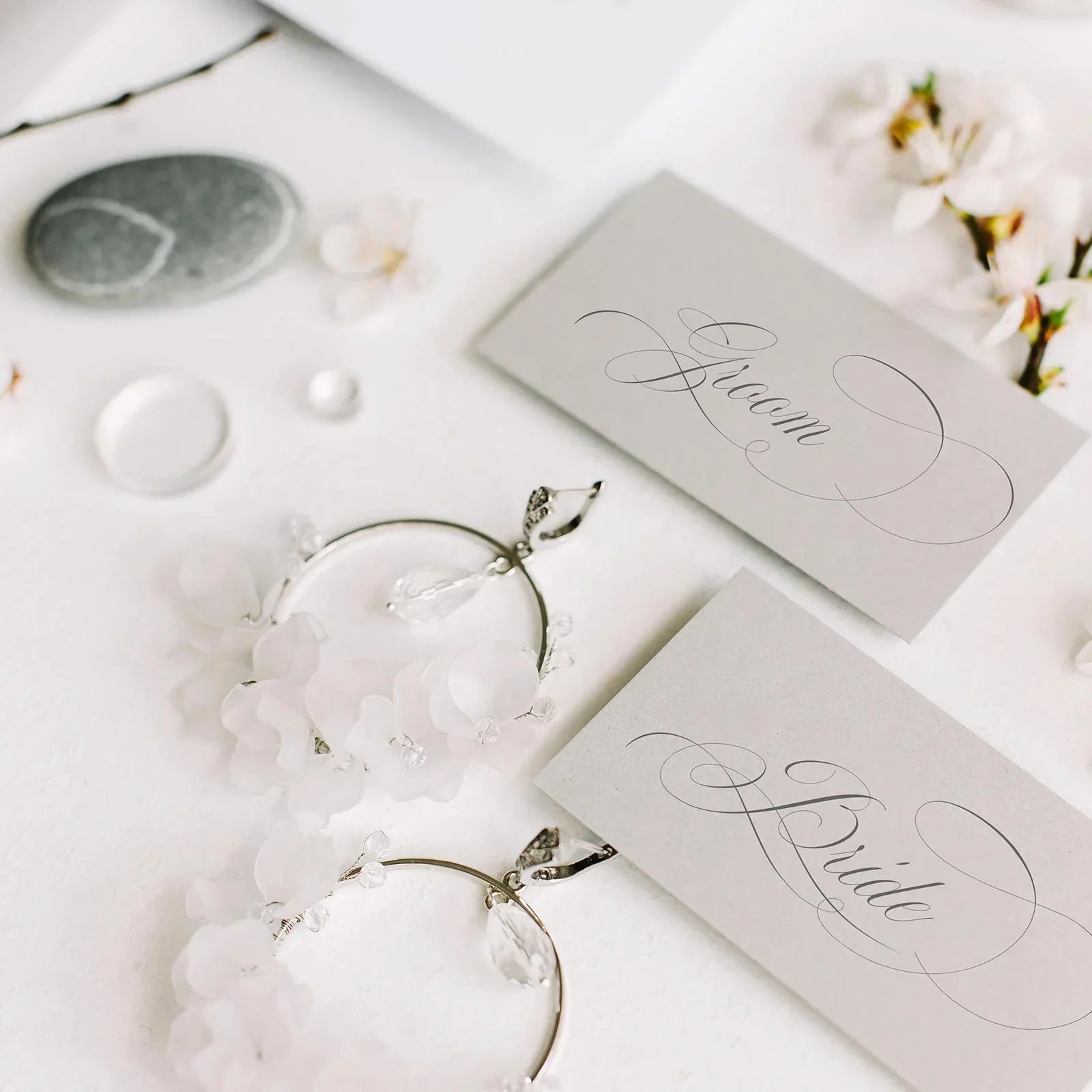 Elegant bride and groom wedding place cards with floral earrings and delicate white blossoms on white surface