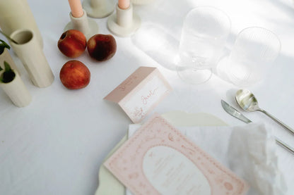 Wedding table setup with folded pink place card labeled Janet, clear glasses, peaches, and cutlery on white linen