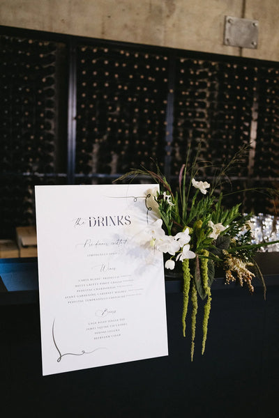 The Drinks elegant wedding bar menu sign with white orchids floral arrangement at wine cellar reception by Felicitations