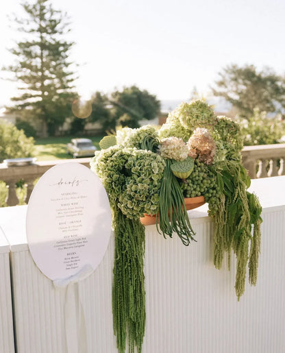 Elegant wedding drinks menu sign with lush green floral arrangement on white bar