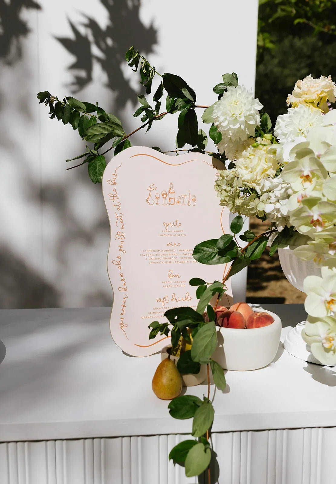 Elegant wedding drinks menu signage with floral arrangement, peaches, and greenery on white table