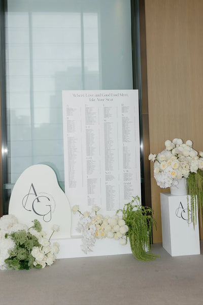Modern wedding seating chart 'Where Love and Good Food Meet' with AG monogram heart-shaped sign and lush white floral arrangements
