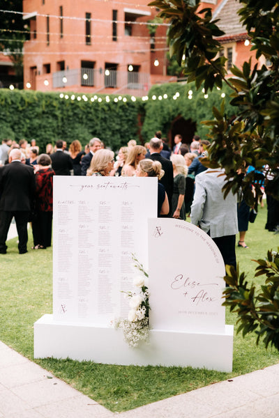 Outdoor wedding seating chart and arch-shaped welcome sign for Elise and Alex with floral details, set in a courtyard with festoon lights