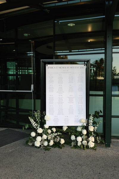 Outdoor wedding seating chart 'Jessica & Nicholas' Hottest 200' on black metal frame, surrounded by lush white roses and baby's breath florals