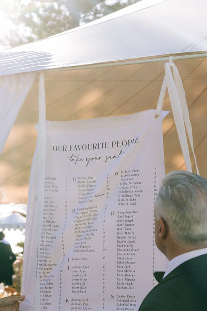 Outdoor wedding seating chart banner with guest names under white tent, man viewing it
