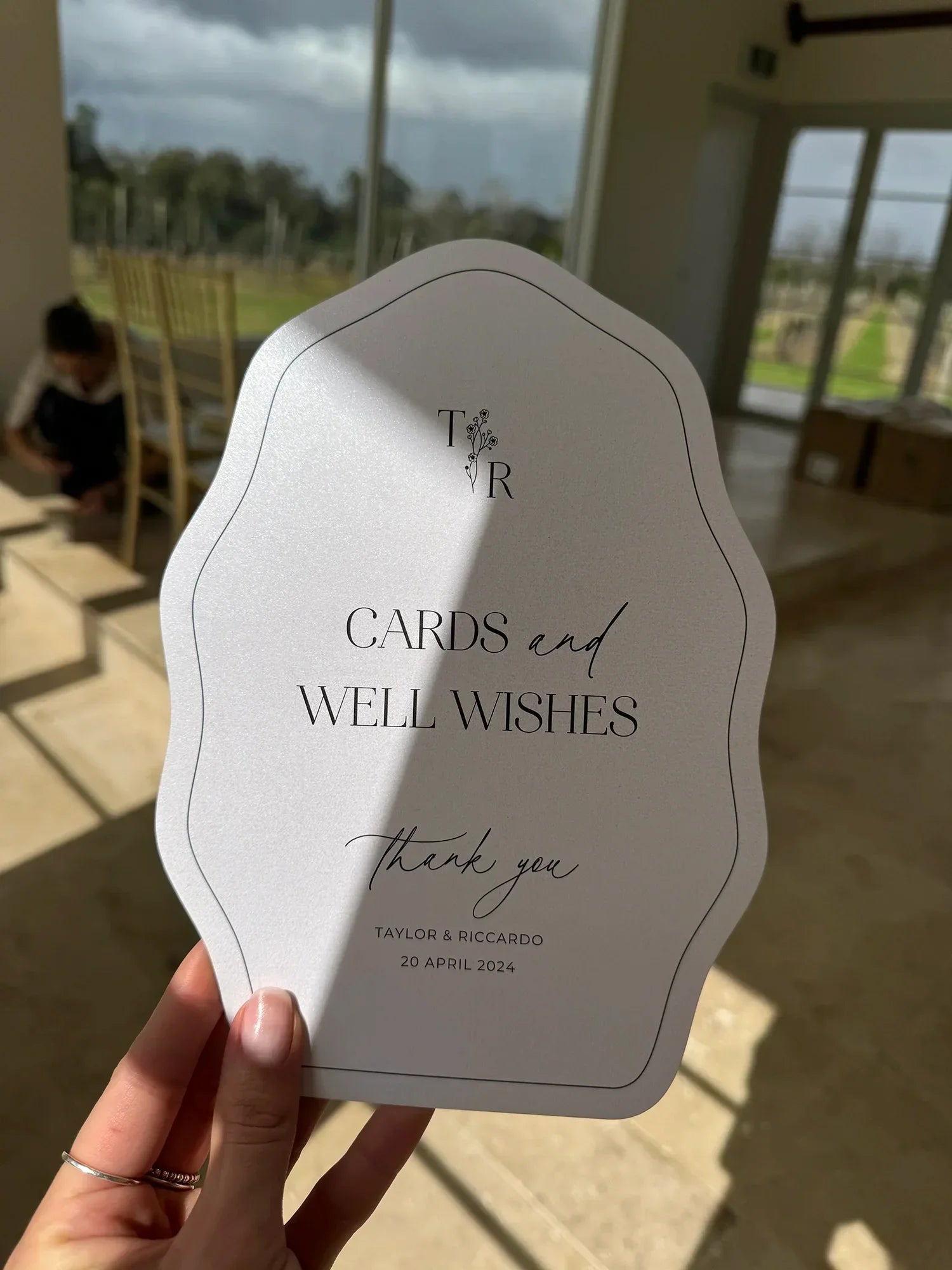 Hand holding elegant wedding card signage reading Cards and Well Wishes, Taylor & Riccardo, 20 April 2024