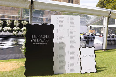 Black wavy-edge wedding seating chart 'The People & Places We Love The Most' beside white Italian benvenuti welcome sign at marquee reception
