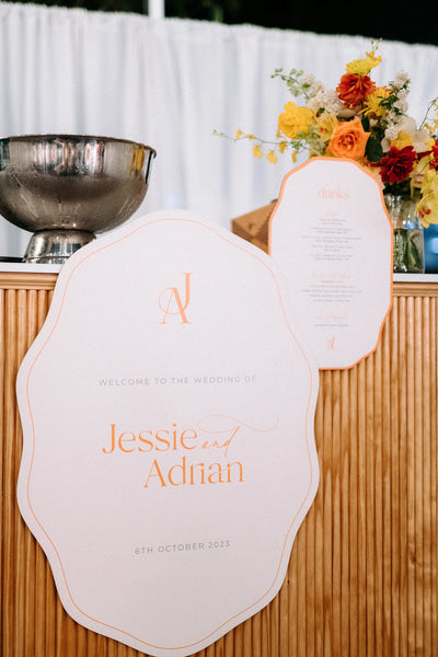 Cloud-shaped welcome sign for Jessie and Adrian in coral with matching drinks menu, styled with bright yellow and orange floral arrangement