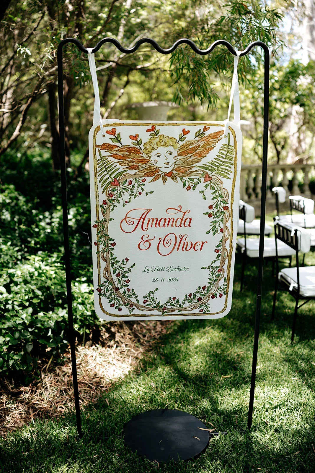 Hand-painted wedding sign for Amanda & Oliver with cherub and floral vine design outdoors