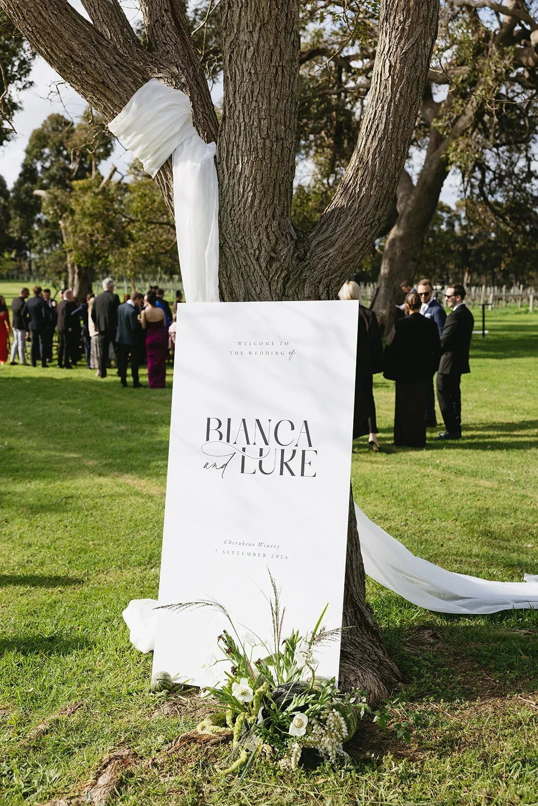 Outdoor wedding welcome sign for Bianca and Luke at Cherubino Winery with floral decor and guests in background