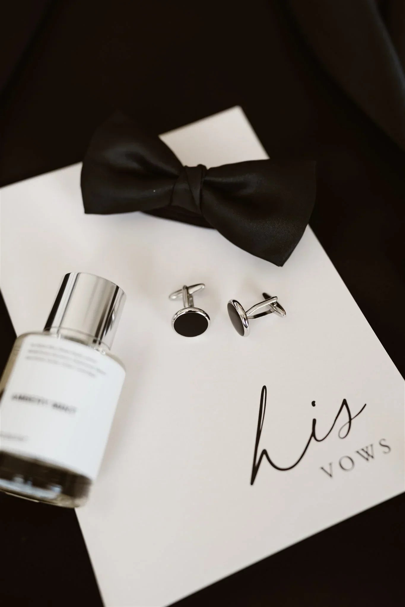 His vows booklet with black bow tie, silver cufflinks, and cologne bottle on black background